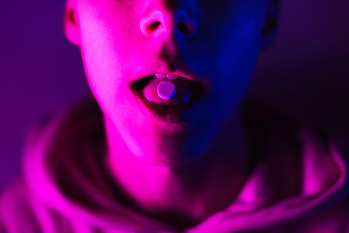 closeup of ecstasy tablet on young person's tongue - pinkish purple lights cast colors on their face - club drugs