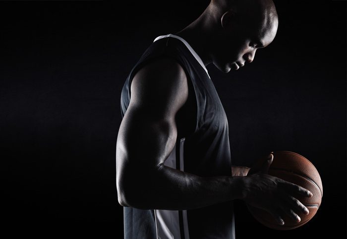 handsome Black male basketball player holding a basketball and looking down, against a black background - athletes and mental health