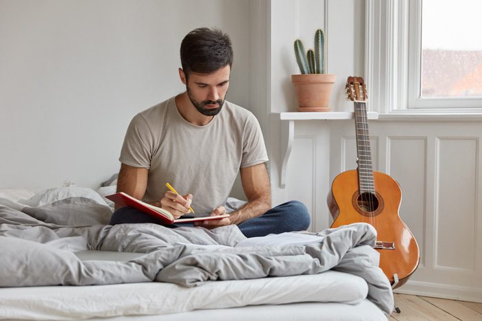 handsome brunette bearded young man sitting cross-legged on his bed while writing in a journal - keeping a journal
