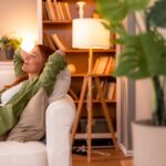 Woman relaxing on a white sofa with her hands behind her head, smiling contentedly in a cozy living room with warm lighting and bookshelves.
