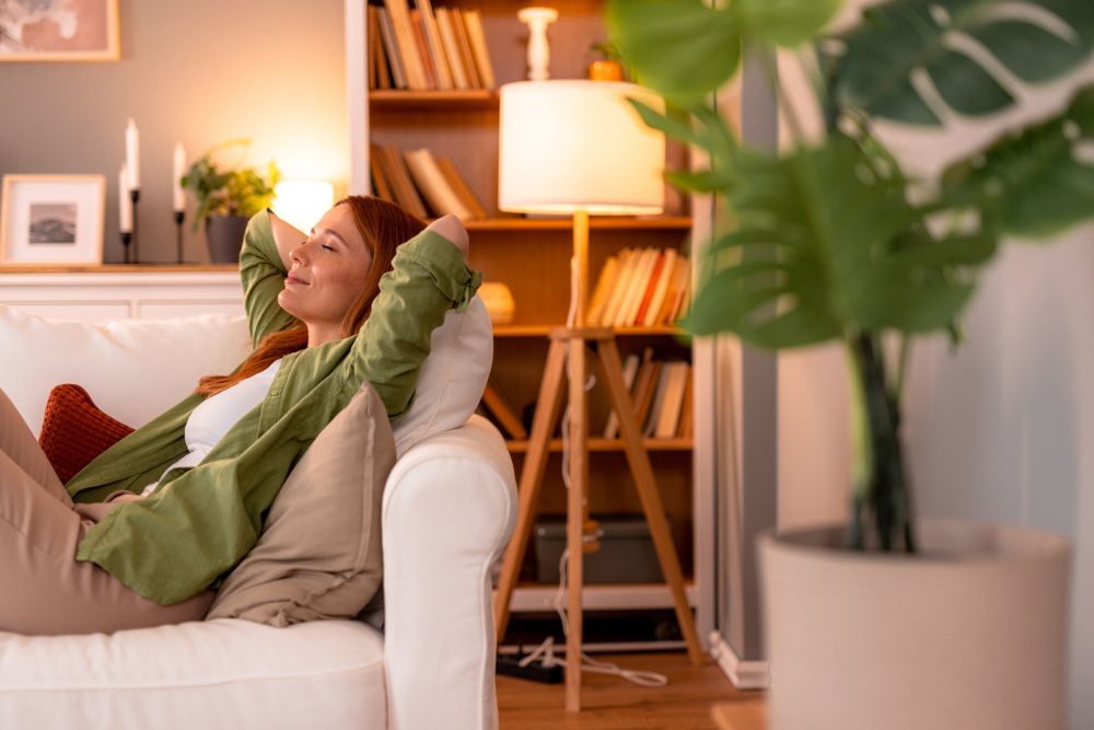 Woman relaxing on a white sofa with her hands behind her head, smiling contentedly in a cozy living room with warm lighting and bookshelves.