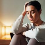 A young woman sits indoors with her eyes closed, resting her head on her hand and looking distressed or exhausted. A warm lamp glows in the background.