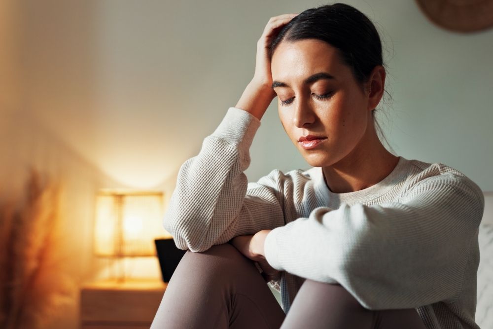 A young woman sits indoors with her eyes closed, resting her head on her hand and looking distressed or exhausted. A warm lamp glows in the background.