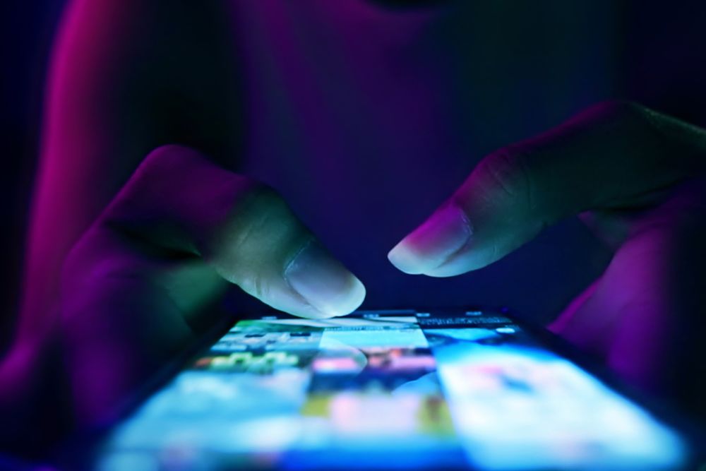 Close-up of a person’s hands scrolling on a smartphone screen glowing with colorful app icons in a dark environment.