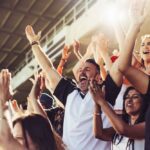 Crowd of sports fans cheering during a match in stadium. Excited people standing with their arms raised, clapping, and yelling to encourage their team to win.