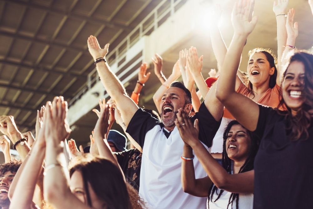 Crowd of sports fans cheering during a match in stadium. Excited people standing with their arms raised, clapping, and yelling to encourage their team to win.