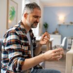 Middle-aged man sitting on a couch in a cozy living room, holding a glass of water and a pill, suggesting he is about to take medication, possibly antidepressants.