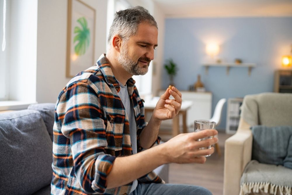 Middle-aged man sitting on a couch in a cozy living room, holding a glass of water and a pill, suggesting he is about to take medication, possibly antidepressants.
