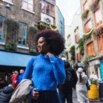 Smiling woman in a blue sweater walking outdoors in a lively urban area with colorful buildings and people in the background.