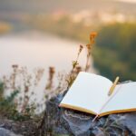 Open journal with a pen resting on it, placed on a rock overlooking a river and nature, symbolizing reflection and mindfulness.