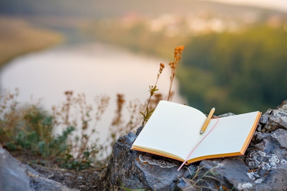 Open journal with a pen resting on it, placed on a rock overlooking a river and nature, symbolizing reflection and mindfulness.