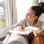 A woman sits by a window writing in a journal, pausing to look outside thoughtfully while holding a pen.