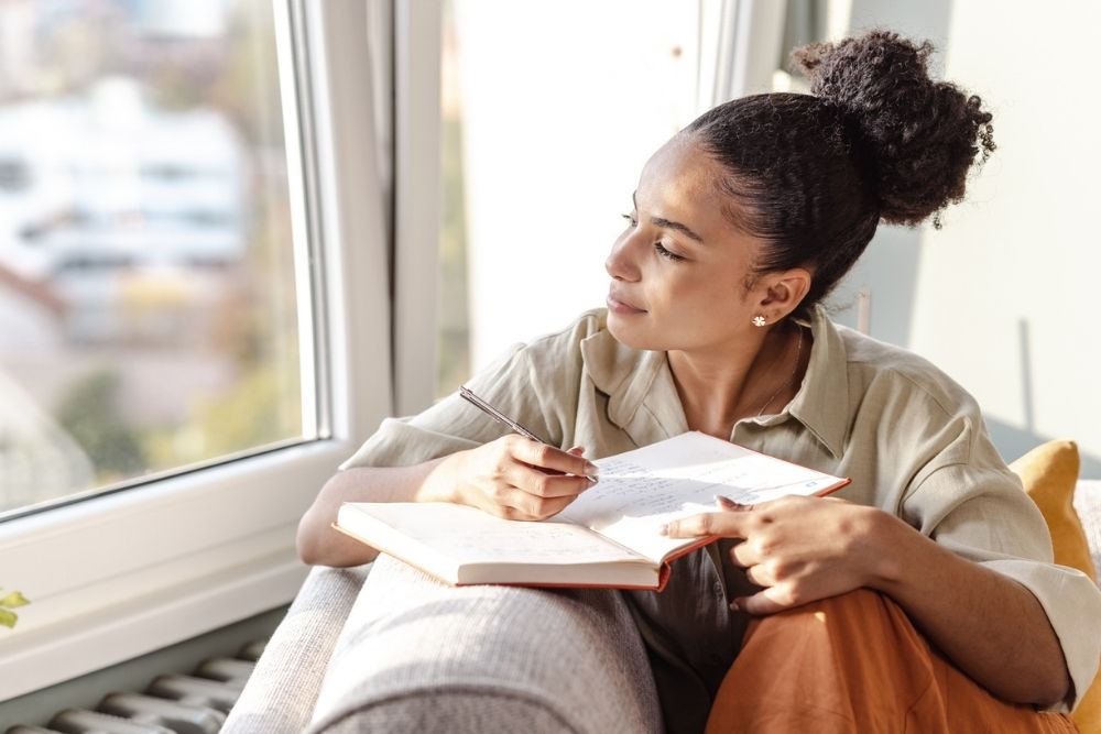 A woman sits by a window writing in a journal, pausing to look outside thoughtfully while holding a pen.