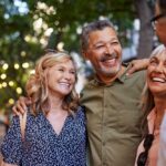 A group of friends standing outdoors, smiling and enjoying each other’s company under string lights in a park or city walkway.