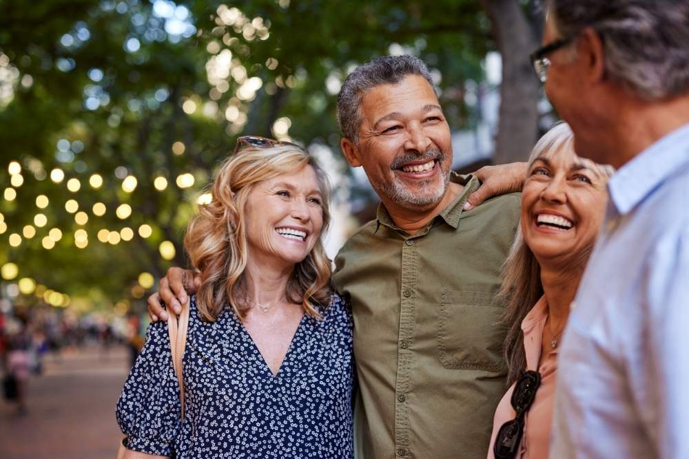 A group of friends standing outdoors, smiling and enjoying each other’s company under string lights in a park or city walkway.