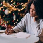 A woman sits at a table near a decorated Christmas tree, writing in a notebook with a warm drink beside her. She looks thoughtful and relaxed as she reflects and writes.