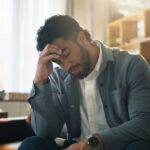 A man sitting indoors with his head resting on his hand, appearing thoughtful or distressed in a quiet, comfortable room.