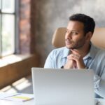A man sits at a desk with a laptop, hands clasped under his chin as he looks thoughtfully out a window in a quiet office setting.