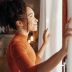 Peak BH - How to Gain Motivation When Depression Makes Everything Feel Hard. A motivated woman looks out a bright window smiling and looking into the distance.